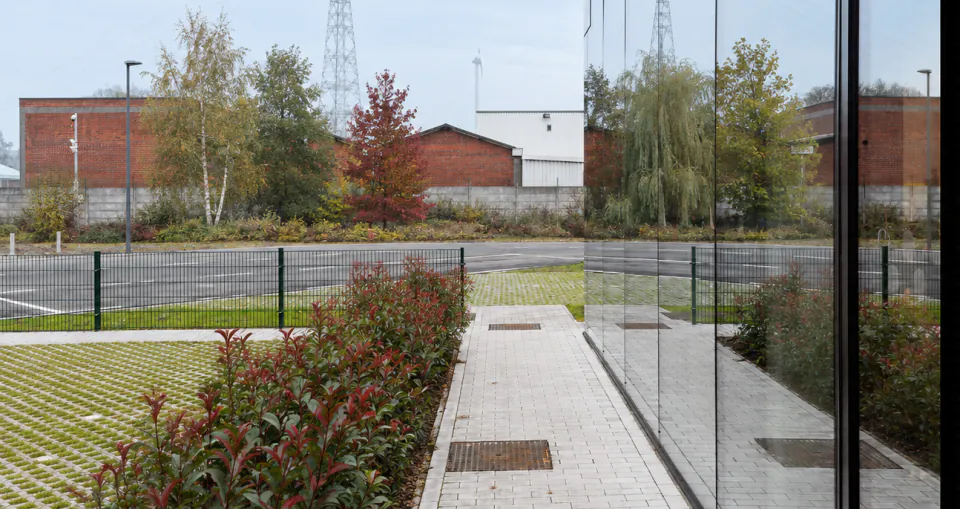 Entrance path beside a glazed building, lined with red-leaved shrubs and permeable paving